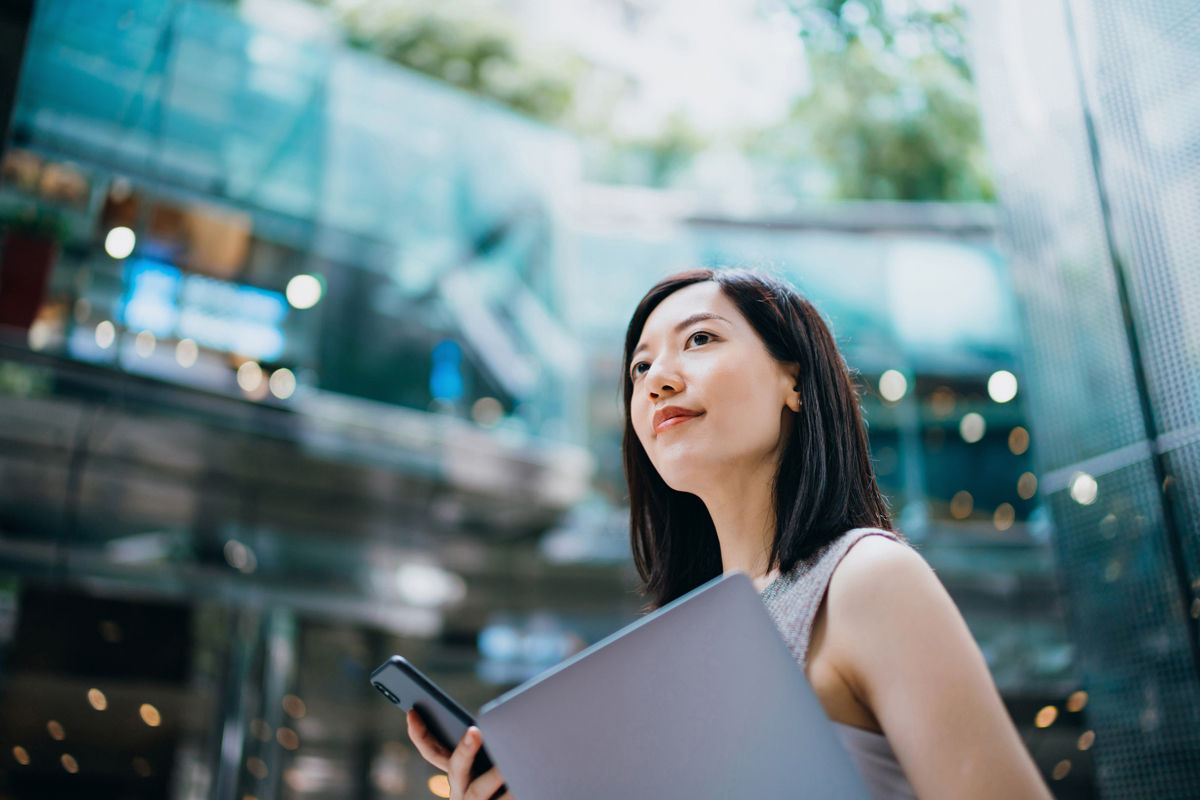 A young woman holding a laptop and phone, looking upward in a modern glass building, symbolizing ambition and personal growth.