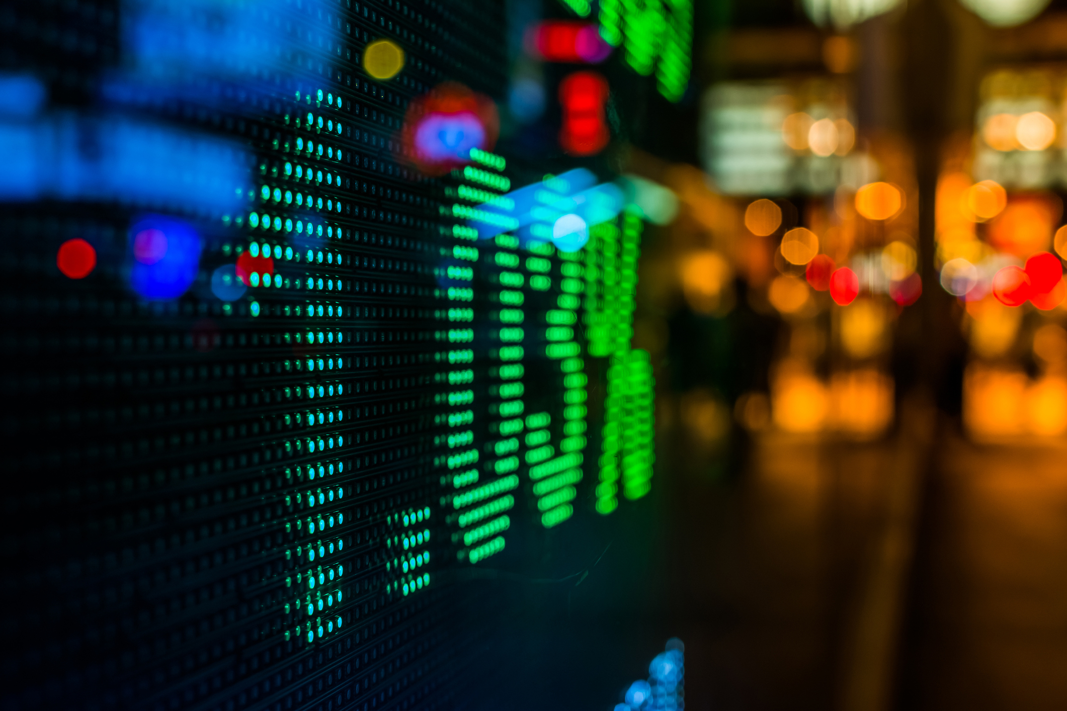Close-up of a digital stock market display showing green numbers and charts, with a blurred city street and lights in the background.