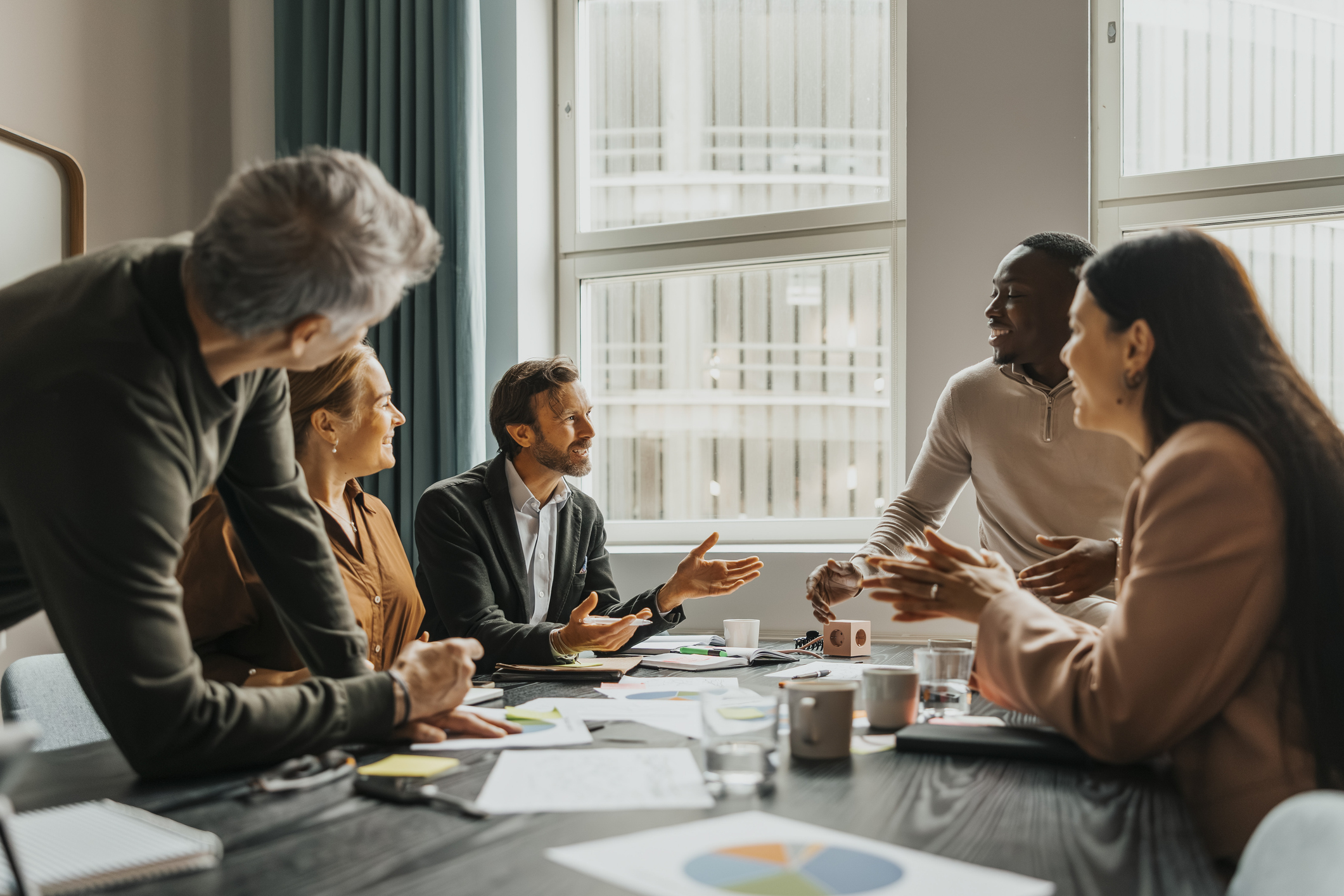  Five professionals collaborate around a conference table in a bright office, discussing documents, charts, and notes during a business meeting.