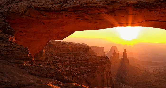 Sunrise over a desert canyon, viewed through a natural rock arch, with glowing orange cliffs and distant stone spires illuminated by golden light.