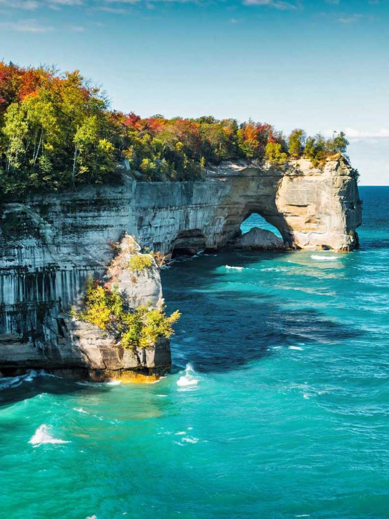 Sandstone cliffs topped with autumn trees above turquoise Great Lakes water.